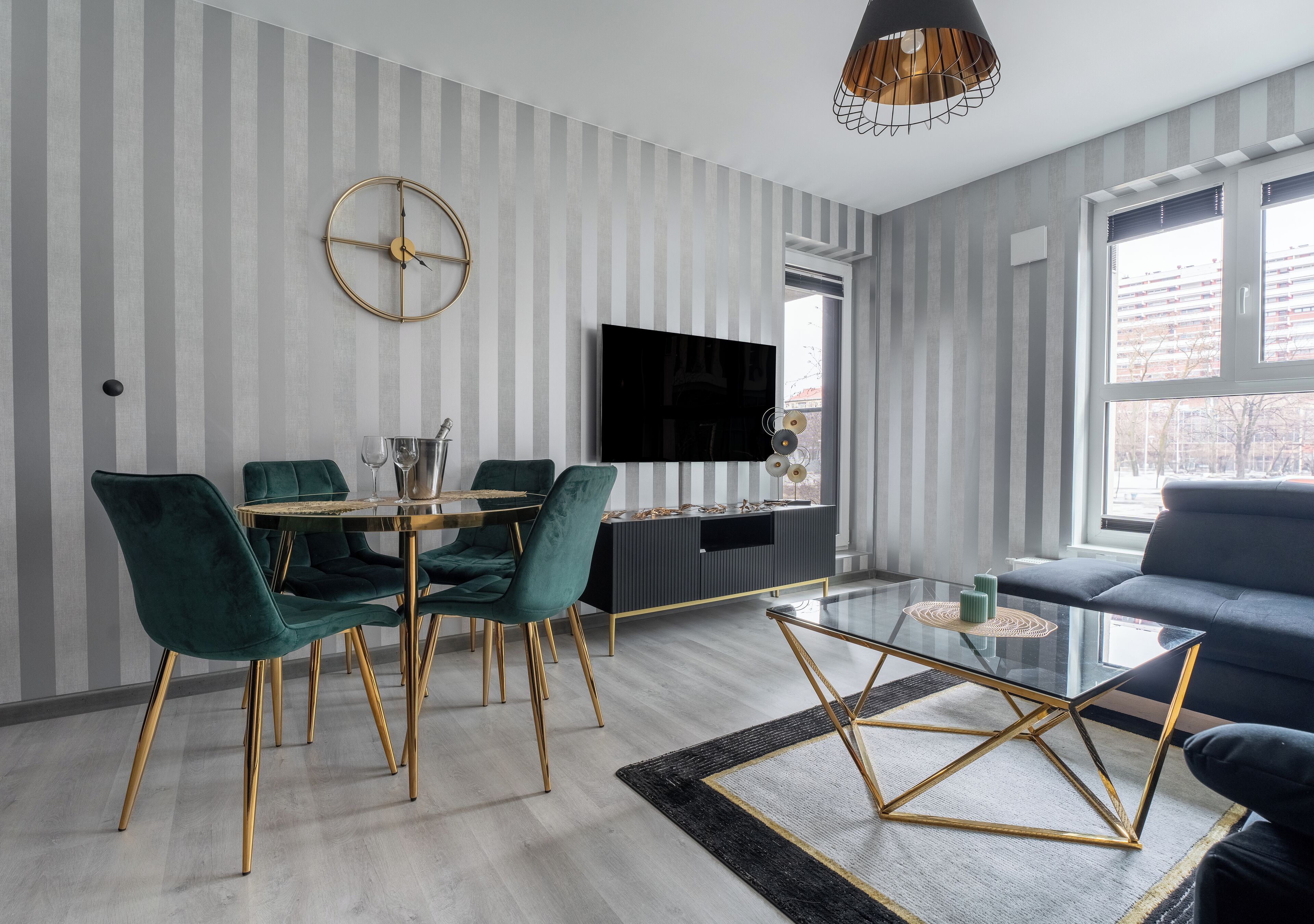 A modern living room with a dining area, featuring striped wallpaper, a round gold clock, a flat-screen TV, and a black sofa.