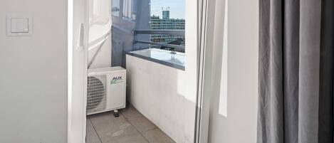 Un pequeño balcón con vistas a un paisaje urbano, una ventana con cortinas y un equipo de aire acondicionado blanco con la etiqueta “AUX DC inverter”.