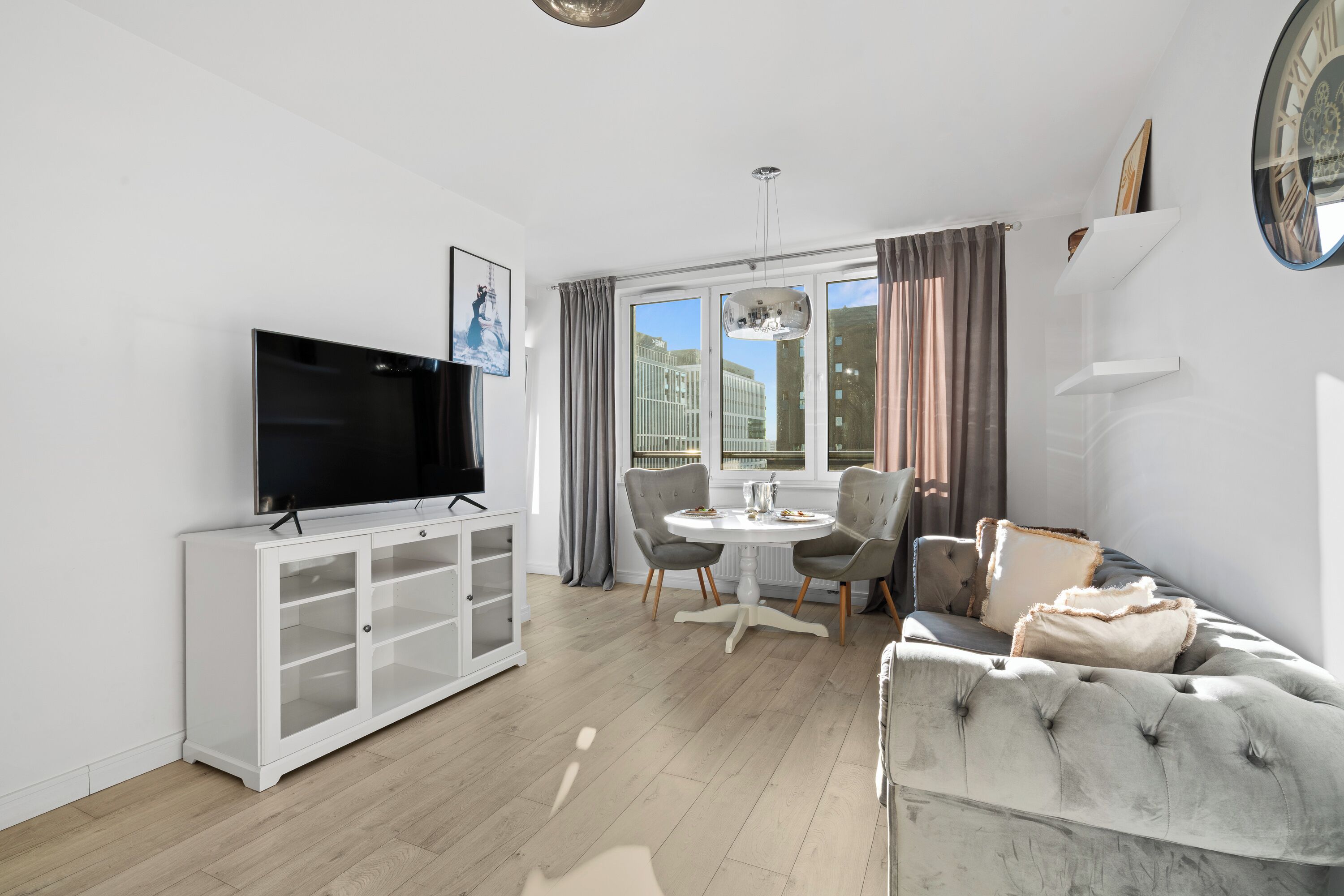 Een moderne woonkamer met een flatscreen televisie, een witte tv-meubel, een grijze bank en een eettafel met stoelen.