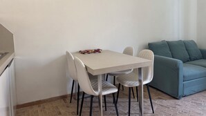 A small, modern living space with a white table and four chairs, a teal sofa, and a patterned bag on the table.