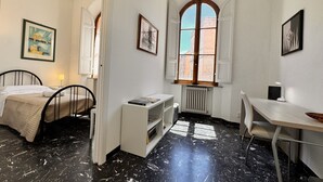 A bedroom with a bed, a desk, a chair, a radiator, and a window with a view of buildings. - Abbadia Panoramic Terrace (Siena)