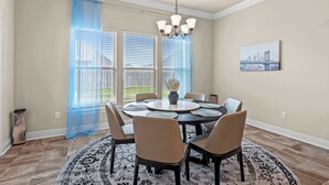 Een eetkamer met een ronde tafel, zes stoelen, een kroonluchter, een vaas en een schilderij aan de muur.