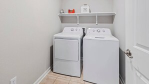 A laundry room with two white washing machines and a shelf above holding a red box and a sign that says "FRESH and CLEAN".