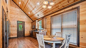 A wooden cabin interior with a dining area, kitchen, and a large window with blinds. - Standing Bear | Romantic Forest Escape for Couples (Broken Bow)