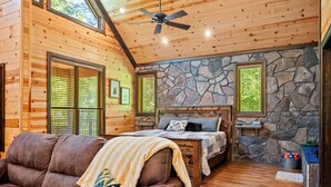 A cozy log cabin bedroom with a stone wall, a bed with a quilt, and a brown leather couch. - Standing Bear | Romantic Forest Escape for Couples (Broken Bow)