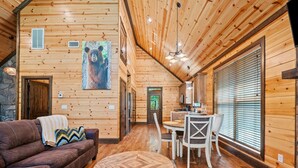 A cozy cabin interior with a wooden ceiling, a dining area with white chairs, a brown leather sofa, and a large bear painting on the wall. - Standing Bear | Romantic Forest Escape for Couples (Broken Bow)