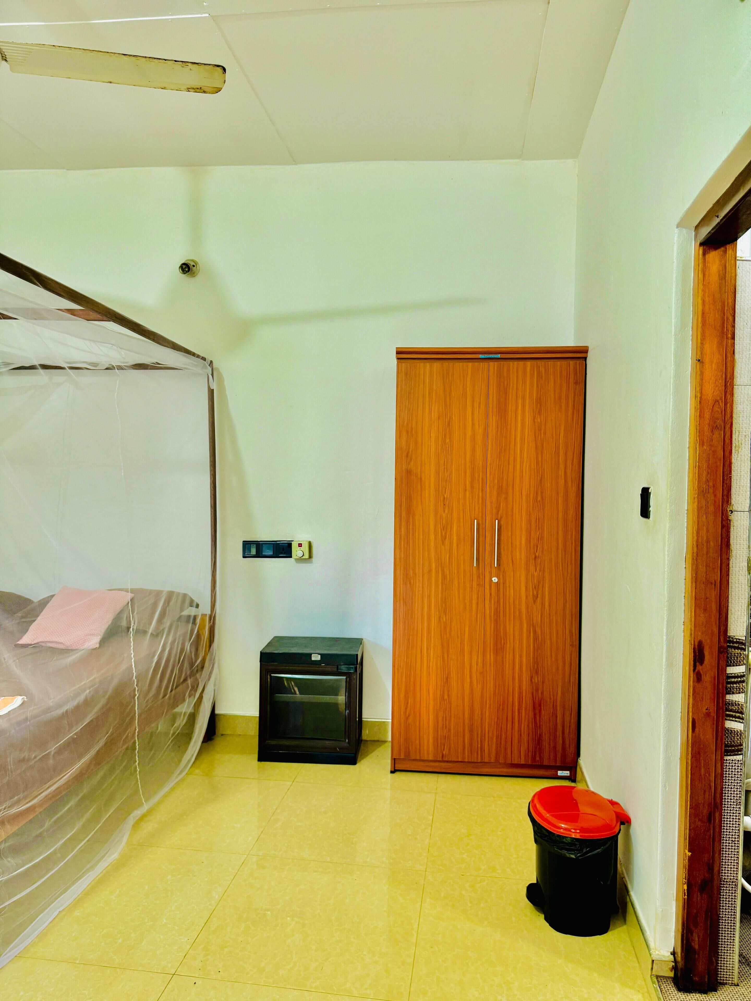 Une chambre avec un lit recouvert d’une moustiquaire, une armoire en bois, un petit radiateur et une poubelle.