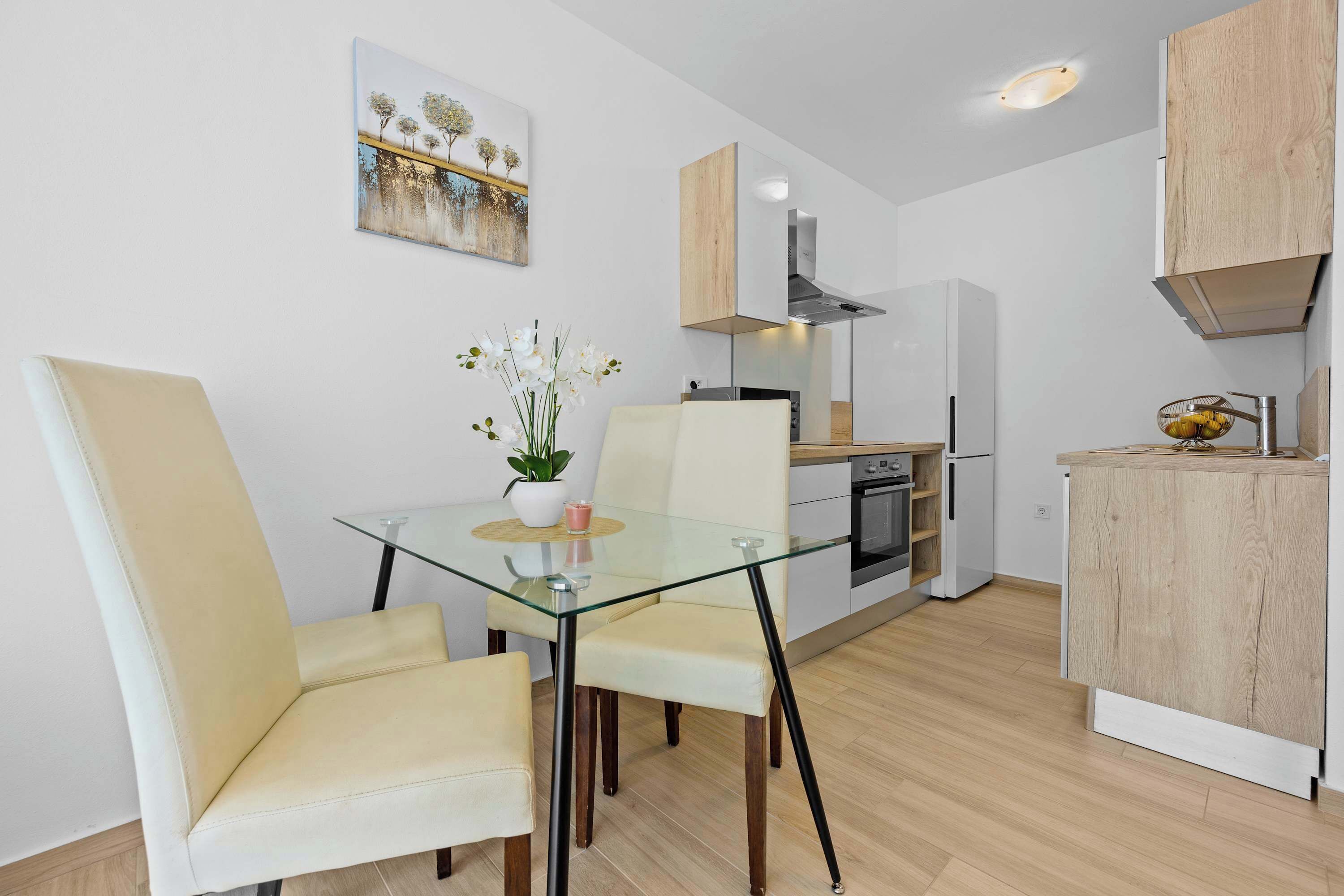 A modern kitchen with a glass dining table, two chairs, a potted plant, and a painting on the wall.