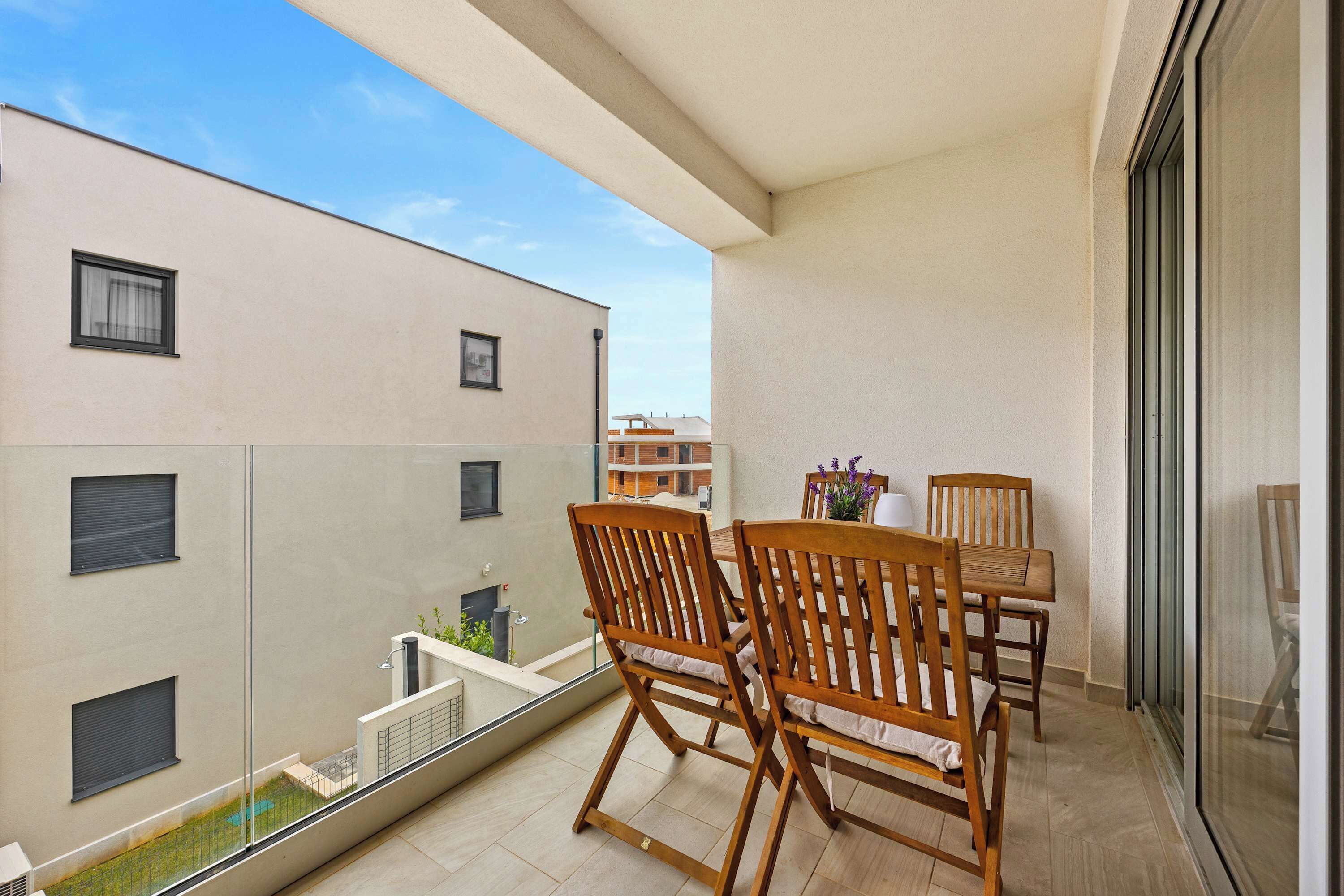 Ein Balkon mit Holzstühlen und einem Tisch, einer Glasreling und Blick auf benachbarte Gebäude.