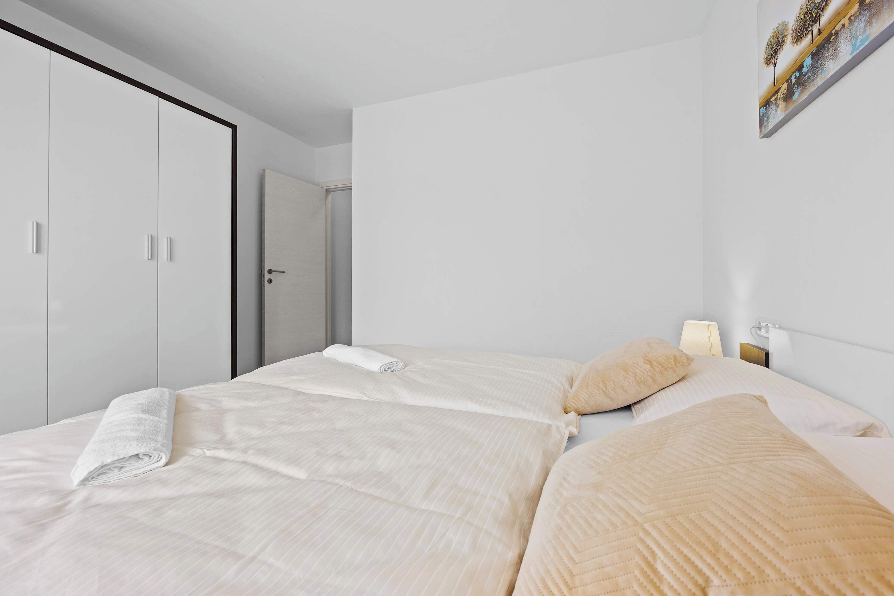 Quarto de hotel com duas camas, um guarda-roupa, um quadro na parede e uma mesa de cabeceira com abajur.