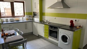 A compact kitchen with a microwave, oven, and washing machine, a dining area with a table and chairs, and a window with a view of a stone wall.
