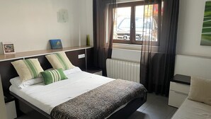 A bedroom with a bed, a nightstand, a dresser, a window with curtains, and a radiator.