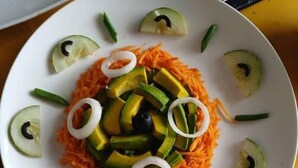 A white plate with shredded carrots, avocado slices, cucumber slices, and green beans arranged on a yellow table.