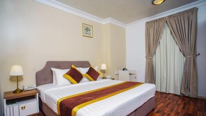 A hotel room with a large bed, two bedside tables with lamps, a nightstand, and a window with curtains.