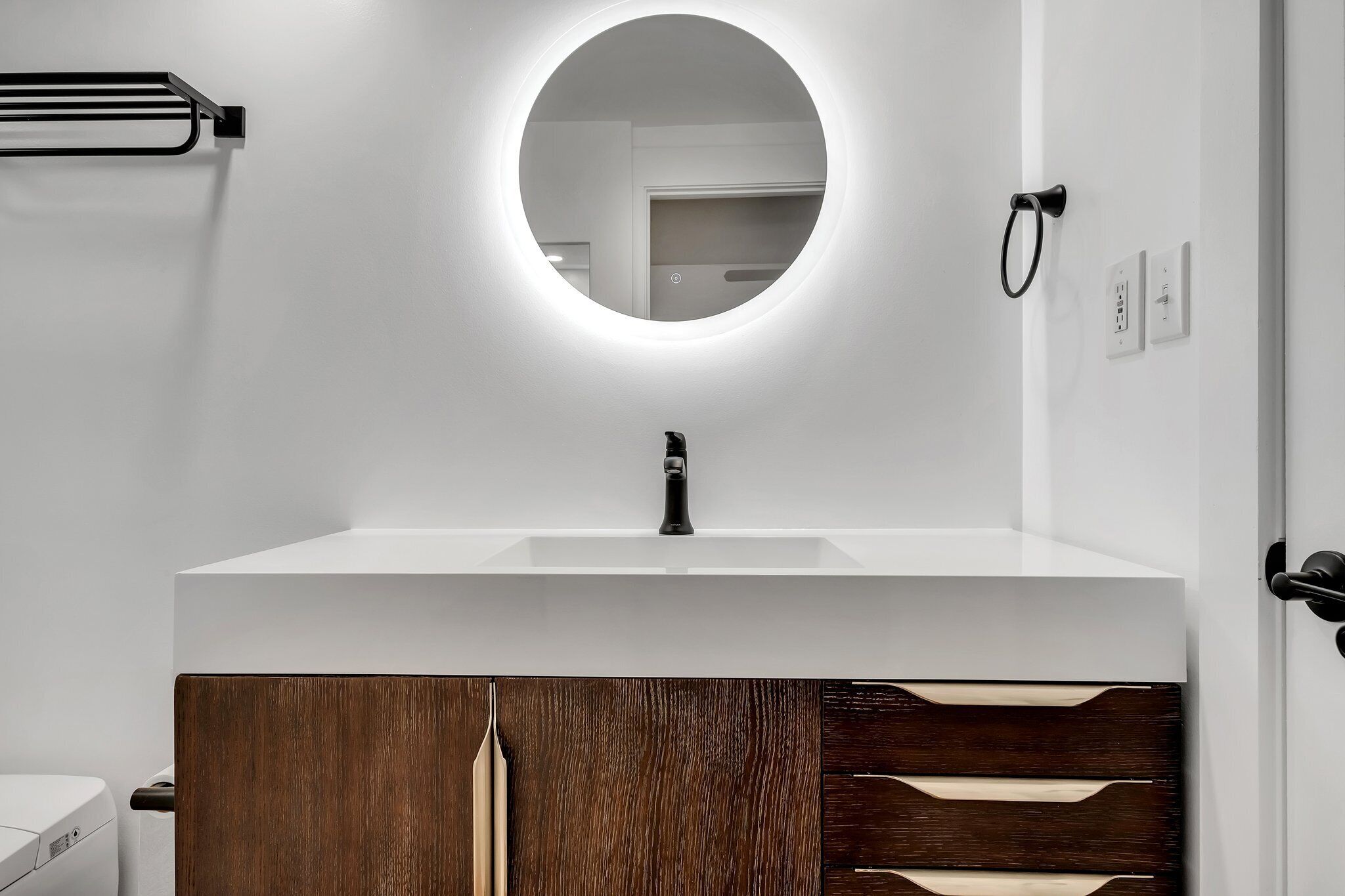 Un baño moderno con un lavamanos blanco, un espejo redondo con luz y muebles de madera.