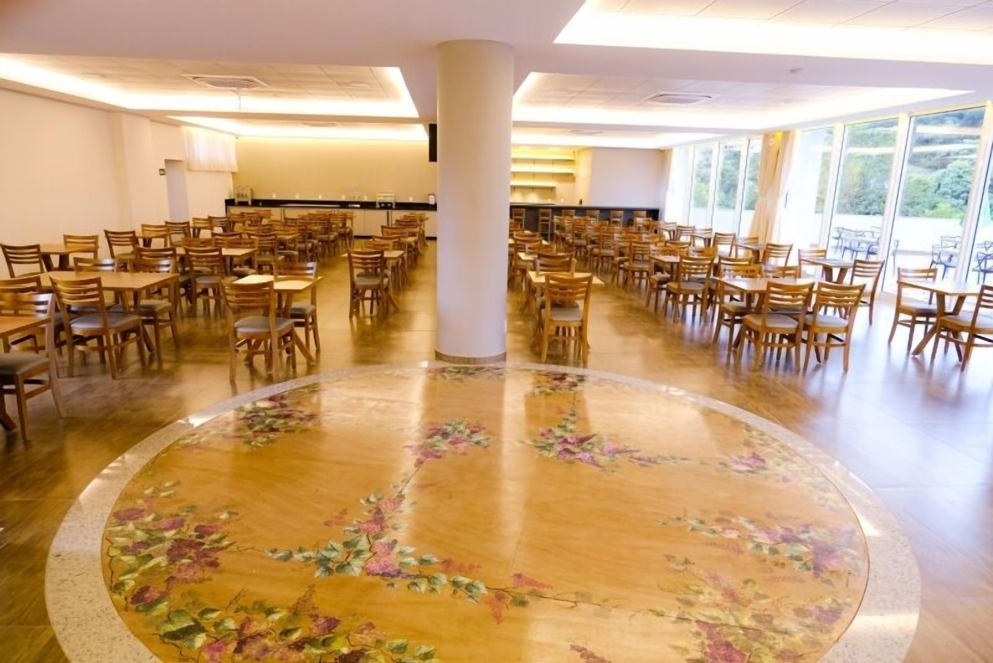 Uma ampla área de jantar com várias mesas e cadeiras, uma mesa redonda decorativa no centro e grandes janelas que proporcionam luz natural.