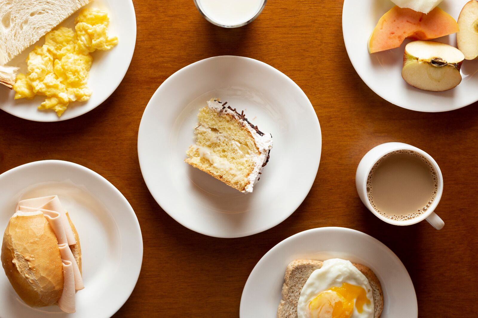 Un desayuno con huevos revueltos, tostadas, jamón, una rebanada de pastel y una taza de café sobre una mesa de madera.