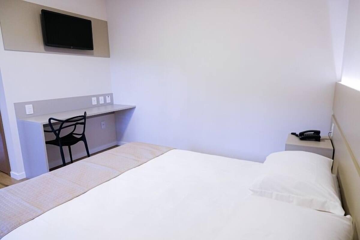A hotel room with a bed, a desk with a chair, a television mounted on the wall, and a bedside table with a phone.