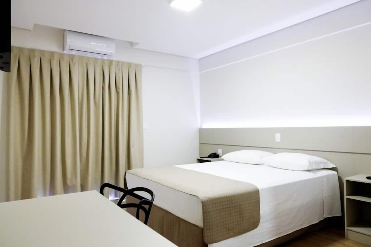 A hotel room with a bed, bedside table, chair, and an air conditioning unit.