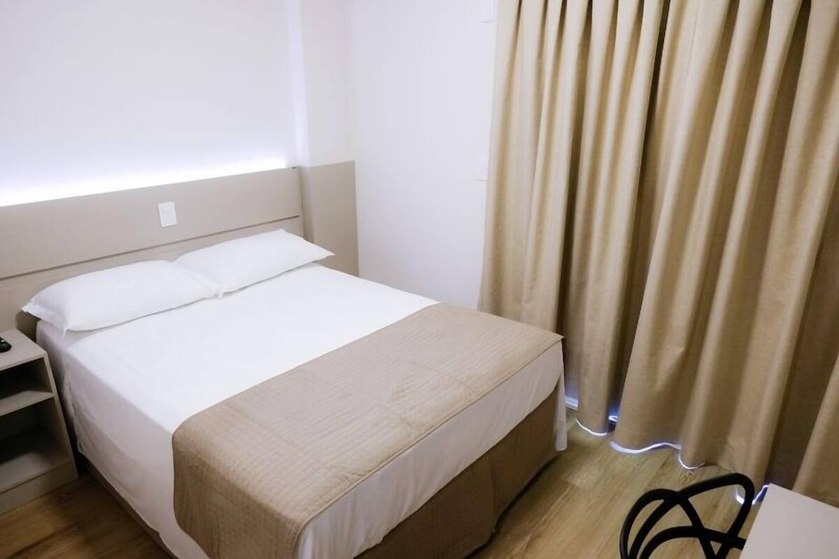 A neatly made bed with a white duvet and a beige throw blanket, a bedside table with a lamp, and a window with beige curtains.
