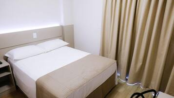 A neatly made bed with a white duvet and a beige throw blanket, a bedside table with a lamp, and a window with beige curtains.