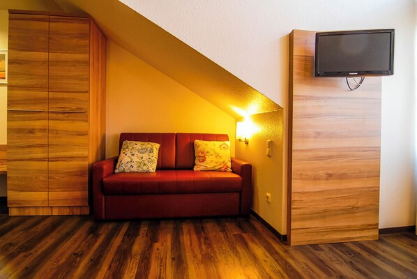 A room with a wooden wardrobe, a red sofa with patterned cushions, and a mounted television. - Adolph*s.hotel (Cologne)