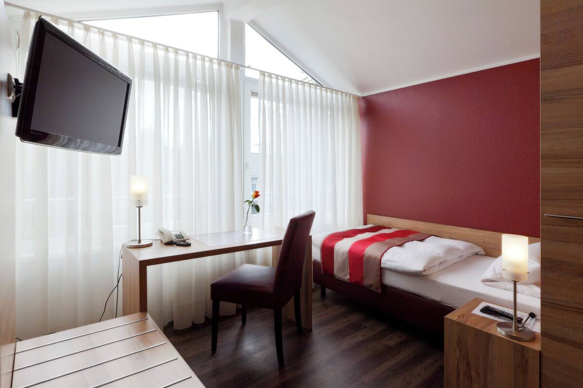 A hotel room with a bed, a desk with a chair, a TV mounted on the wall, and a bedside table with a lamp.