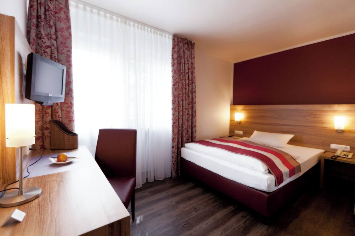 A hotel room with a bed, a desk with a lamp, a television, and a nightstand with a phone.