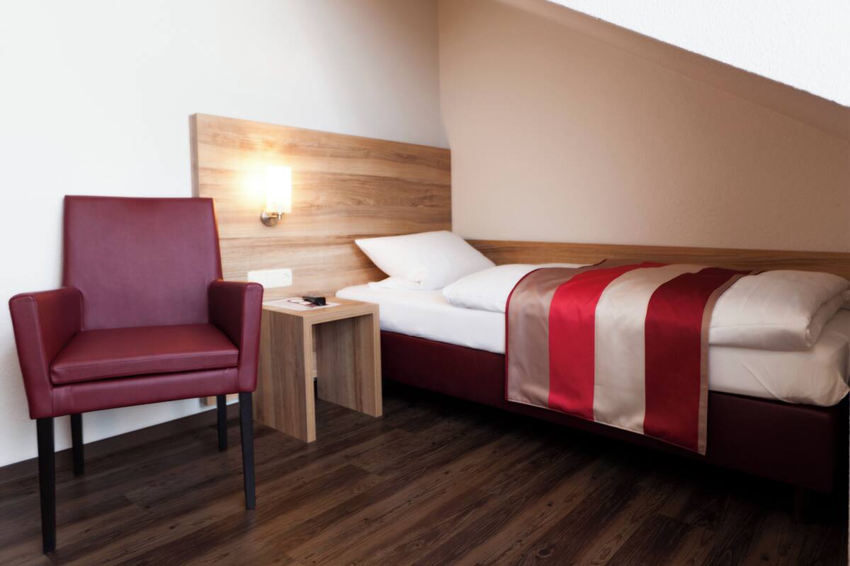 A hotel room with a single bed, a red chair, a small wooden nightstand, and a wall-mounted light.