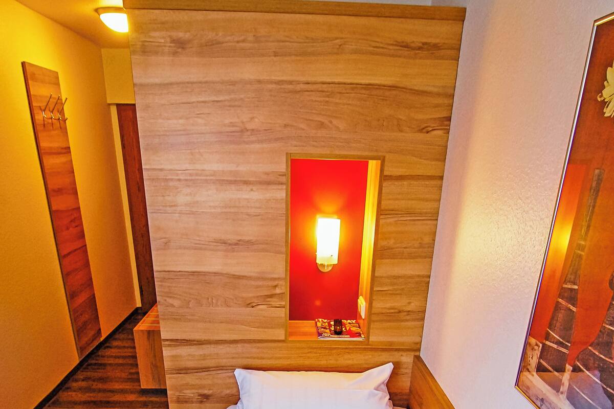 A hotel room with a wooden headboard, a bed with white and red bedding, and a wall-mounted light fixture.
