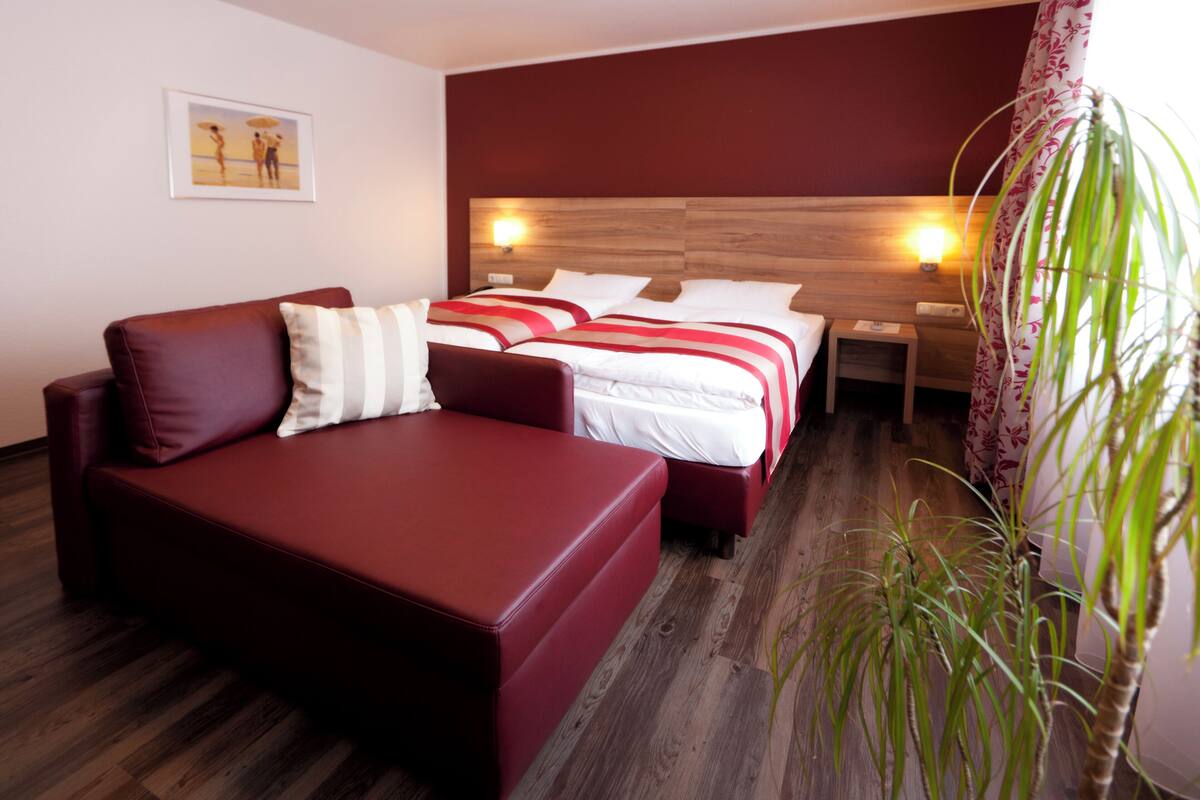 A hotel room with a red sofa, two beds with white bedding and red stripes, wooden headboard, bedside lamps, and a potted plant.