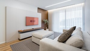 A modern living room with a flat-screen TV mounted on a white wall, a beige sofa, a wooden coffee table, and a large window with sheer curtains. - GuestReady - Beach and Pool Escape in Gaia (Vila Nova de Gaia)
