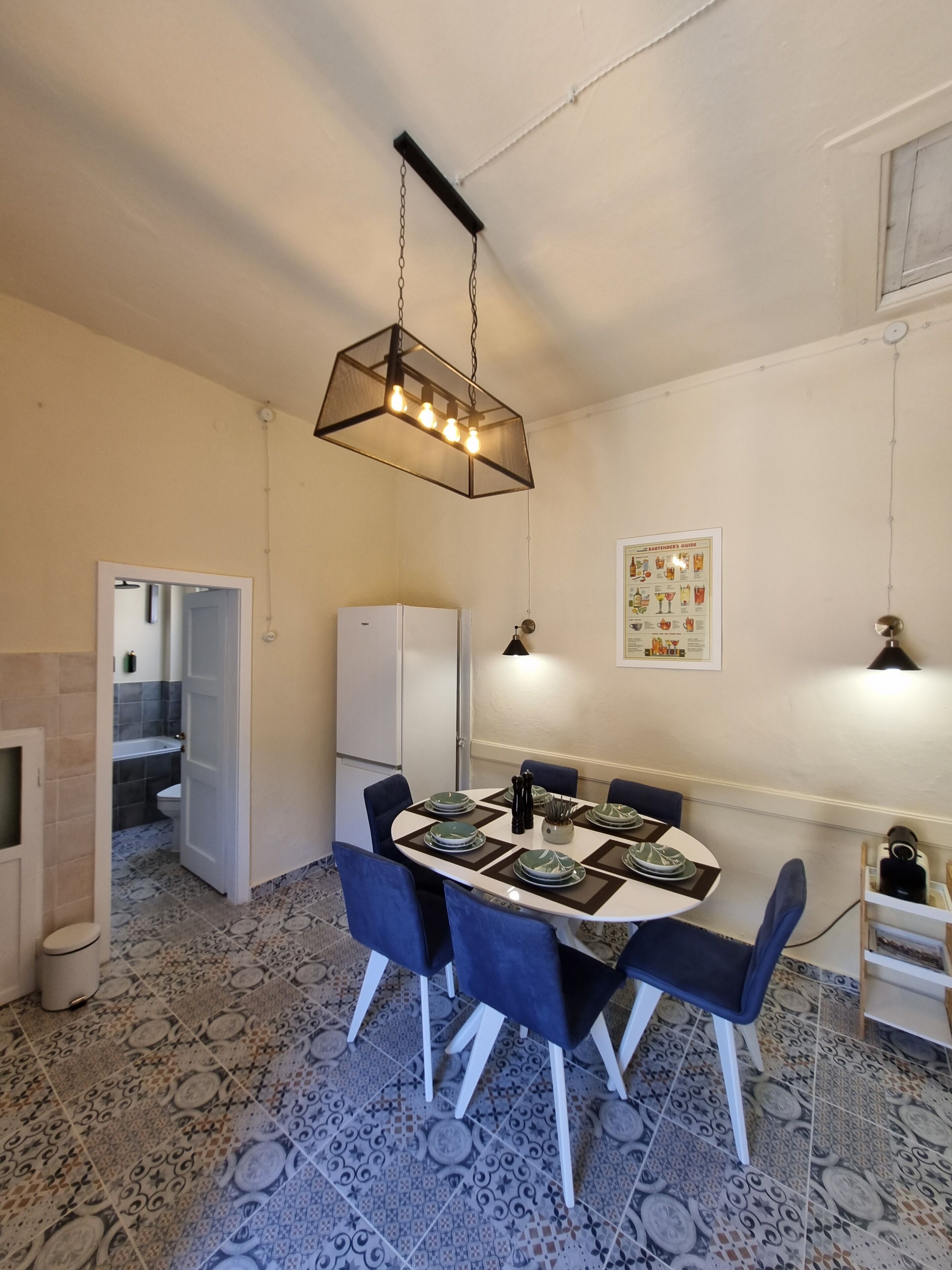 A dining area with a round table, blue chairs, patterned floor tiles, a hanging light fixture, and a refrigerator in the background.
