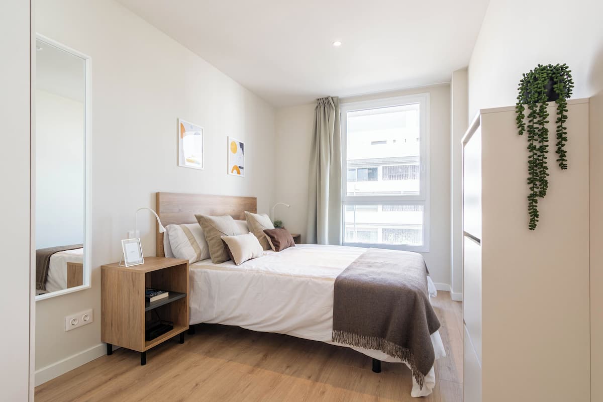 A modern bedroom with a large bed, bedside tables, a mirror, and a window with a view of buildings.
