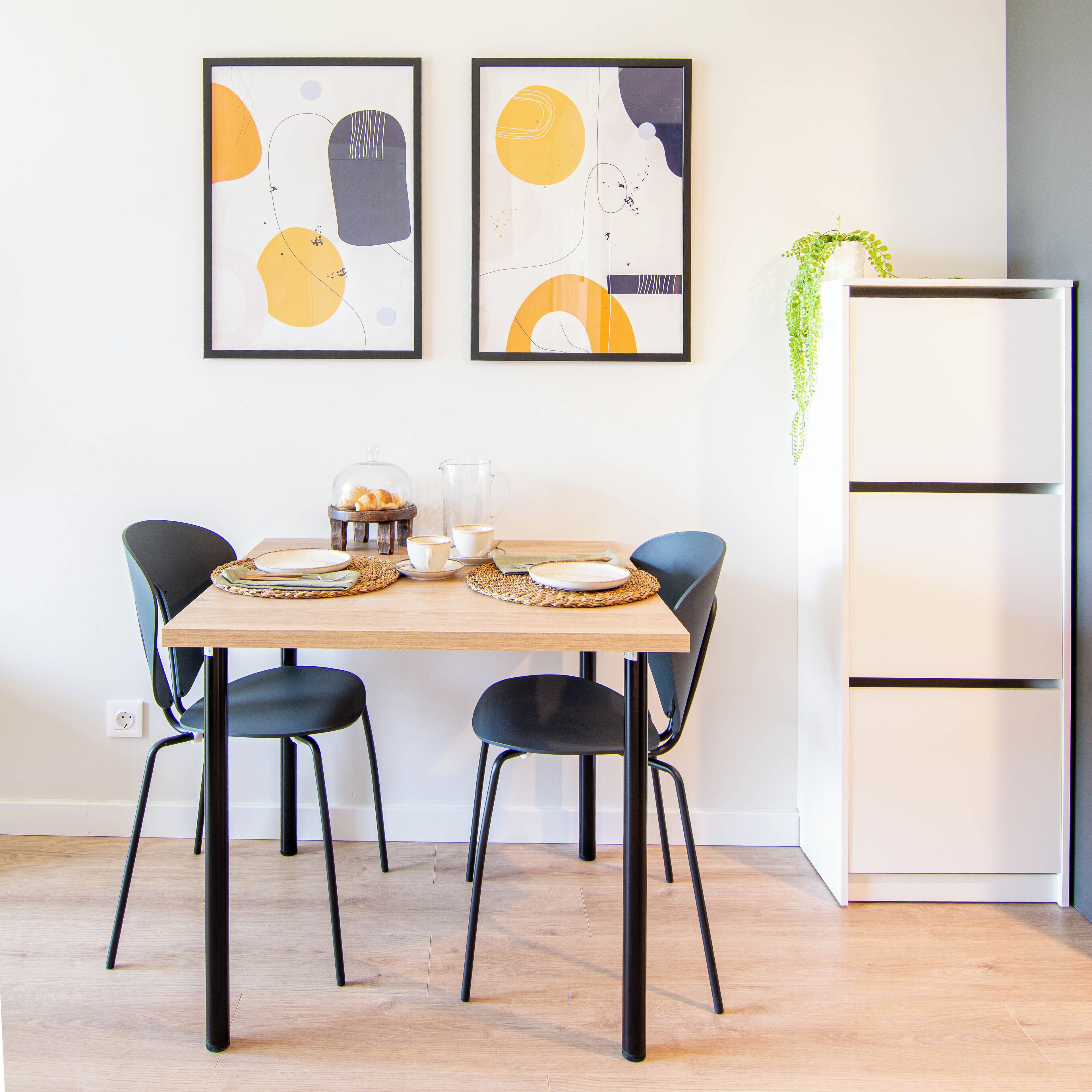A dining area with a wooden table set for two, black chairs, and two abstract paintings on the wall.