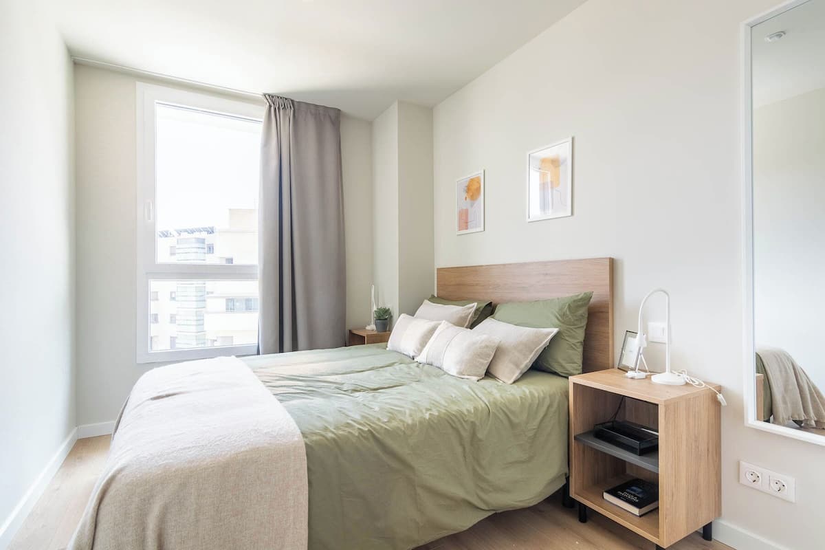 A bedroom with a bed, a nightstand, a bedside table, a mirror, and a window with curtains.