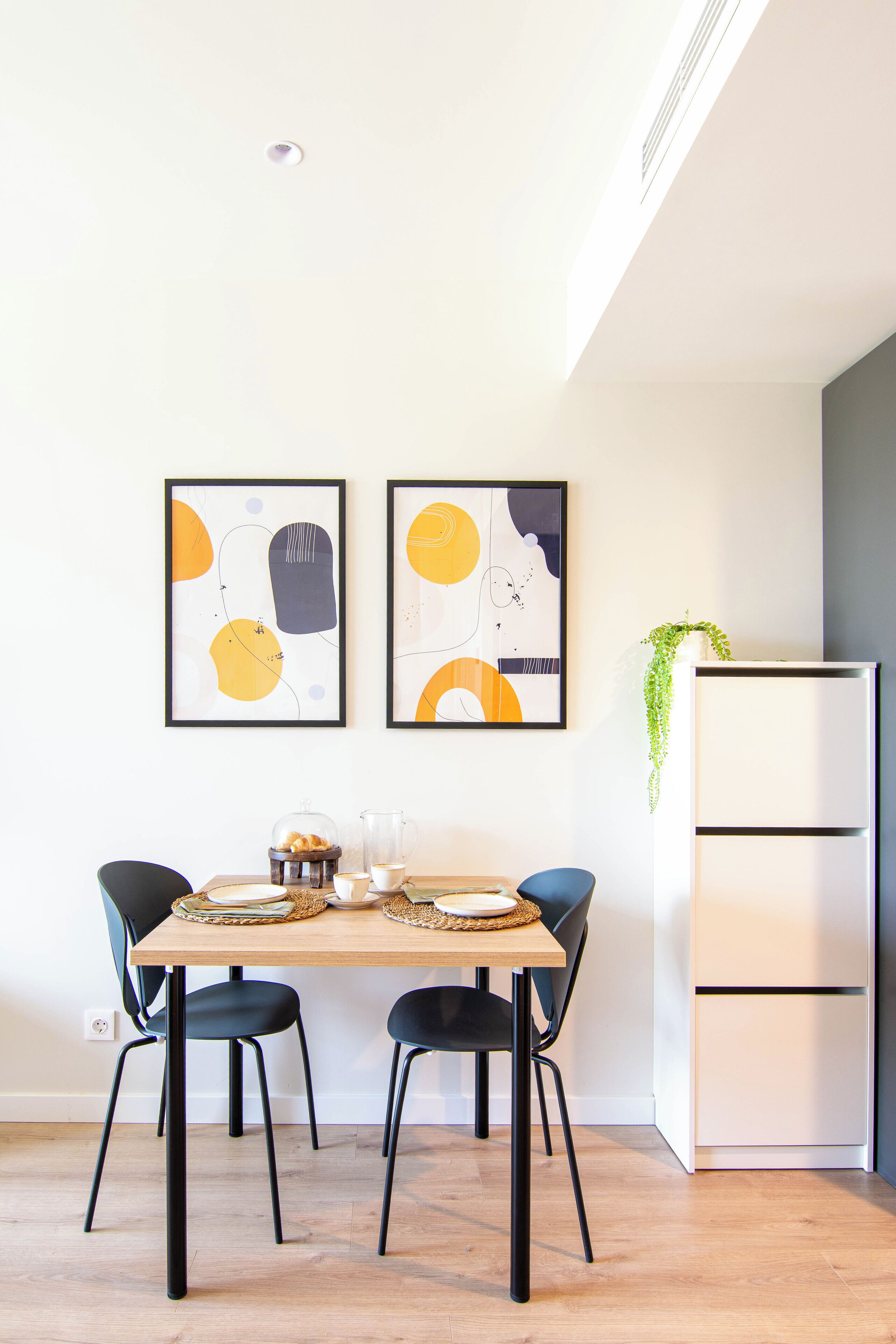 Un comedor con una mesa de madera, sillas negras y dos cuadros abstractos en la pared.