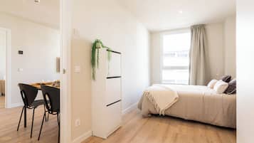 Un dormitorio moderno con una cama, un escritorio con sillas y una planta pequeña.