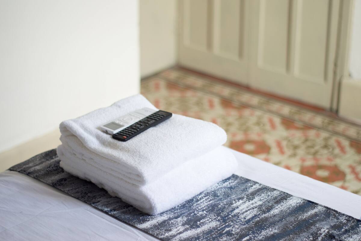 A neatly folded white towel with a remote control on top, placed on a patterned mat beside a bed.