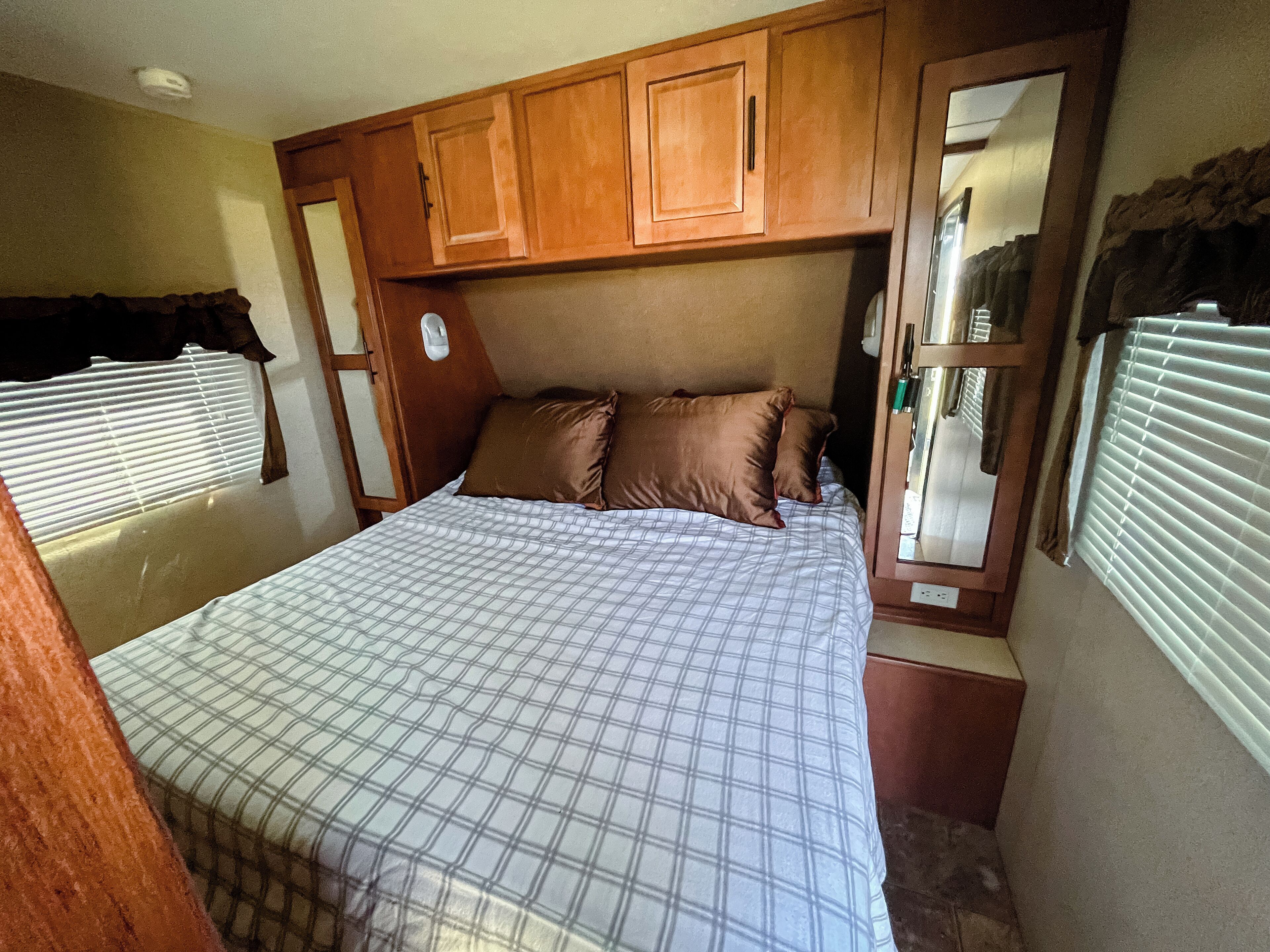 Une chambre compacte dans un camping-car, comprenant un lit, une tête de lit en bois et une fenêtre avec des stores.