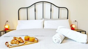 Un lit bien fait, avec une tête de lit en métal, des oreillers blancs et une serviette enroulée. On peut voir une tablette avec des croissants et du jus d’orange posée sur le lit. Deux lampes de chevet aux abat-jour rouges sont visibles.