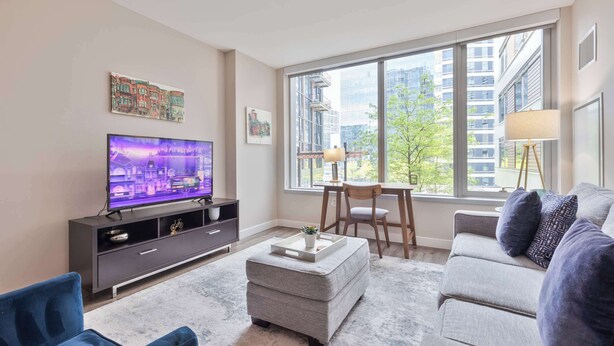 A modern living room with a flat-screen TV, a grey sofa, a coffee table, and a window with a city view.