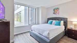A modern bedroom with a large window, a bed with white bedding and blue pillows, a dark headboard, and a nightstand with a lamp.