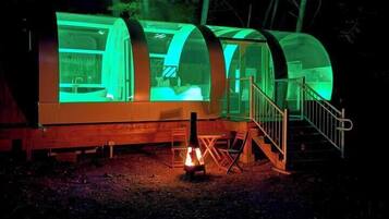 Een moderne, cilindervormige boomhut met een glazen omheining, een klein terras en een trap naar de ingang.