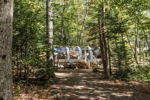 Une zone boisée abritant un camping-caravaning avec un espace couvert comprenant une table et des chaises, un barbecue et un escalier menant à une terrasse.