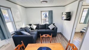 Un espacio habitable pequeño con una mesa de comedor de madera, cuatro sillas, un sofá y un televisor fijado a la pared.