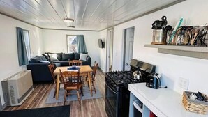 Cocina compacta con estufa, lavamanos y una pequeña encimera. Hay un rincón comedor con una mesa y sillas, y un sofá al fondo.