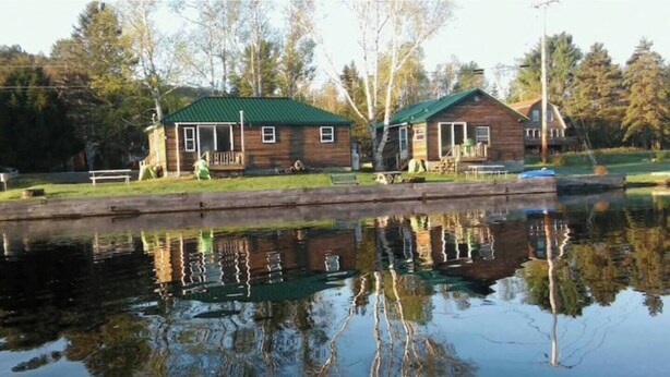 Dos cabañas de madera con techos verdes, un muelle y un lago tranquilo que refleja las construcciones.