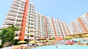 A high-rise hotel with multiple balconies, a swimming pool area with sun loungers and umbrellas, and palm trees. - Praia da Rocha River View 740 (Portimão)