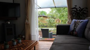 A living room with a sofa, a coffee table with plants, a TV, and a view of a house and greenery outside. - Palmocolodge Lodge - Jocomo Parc a nice family campground with many facilities (Lanaken)